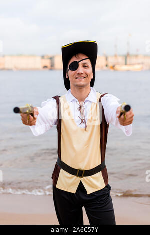 Smiling man in a pirate costume pointed two toy guns into the lens on the sandy beach. Stock Photo