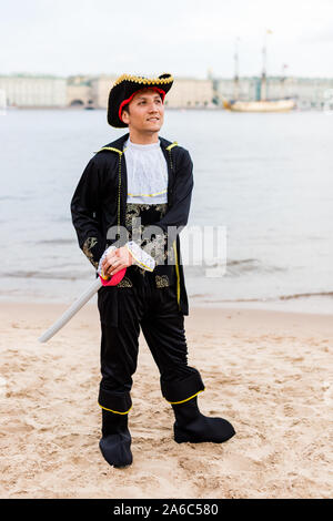 Young Caucasian man in black and white pirate costume holding toy sword and looking away on the sandy beach. Vertical. Stock Photo