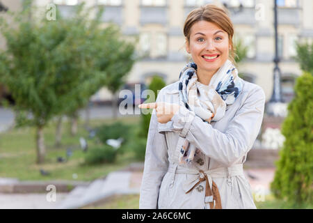 Middle-aged woman points a finger to the side while standing against the backdrop of the city and the park. Stock Photo