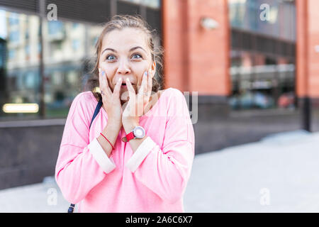 Joyful Caucasian young hipster woman in pink blouse putting hands on cheeks in delight. Positive emotions concept. Stock Photo