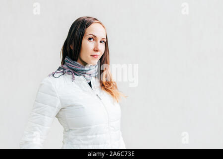 Young Caucasian woman in white jacket with long brunette hair looks into the lens with little smile on background solid monochrome wall. Stock Photo