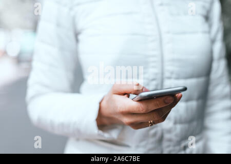 Close up of woman's hand in white jacket holding grey smartphone and scrolling site on the street. The side of the phone. Stock Photo