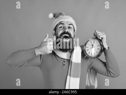 Santa holds old clock showing thumbs up. Winter holiday and countdown concept. Man with beard and cheerful face celebrates Christmas. Santa Claus waits for New Year on red background Stock Photo