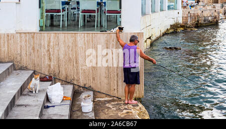 Maltese fisherman patting stray calico cat while catching the fish with rod in another hand. Red cat is sitting on steps waiting for fresh fish Stock Photo