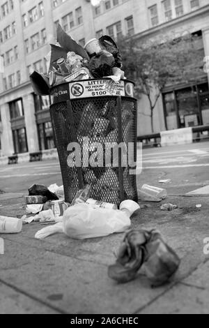 rash can overflowing on urban sidewalk.  Public waste bin full with plastic and paper bags and containers in street.  No people Stock Photo