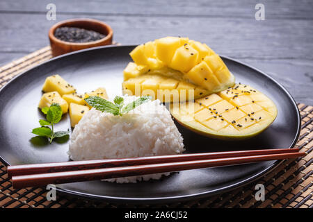 close-up of thai dessert, creamy Sticky Rice cooked with coconut milk and served with fresh Mango on a black plate with chopsticks Stock Photo