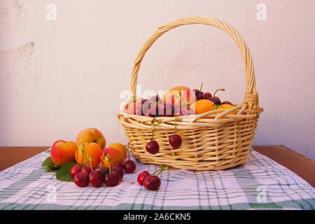 A basket with summer fruits such as cherries, peaches and apricots on a kitchen towel and rough white background Stock Photo