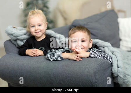 Little girl and boy lying t on the couch covered with a gray knitted blanket and smiling. Brother and sister grimacing and fooling around on the backg Stock Photo