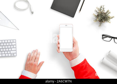 Santa holding smart phone with isolated screen for mockup. Santa's office desk with papers, keyboard, folder, charger cable... Top view, flat lay. Stock Photo