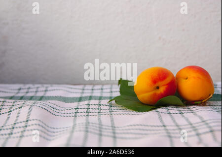 To the right of the image there are two apricots and some green leaves leaving room for texts in the center of a rough white background Stock Photo