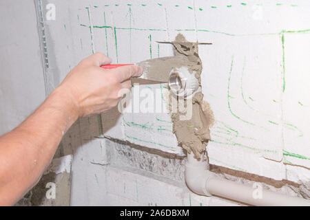 Builder holds a spatula in his hand and covered with cement mortar hole with pipe in the wall with lens flare. Stock Photo