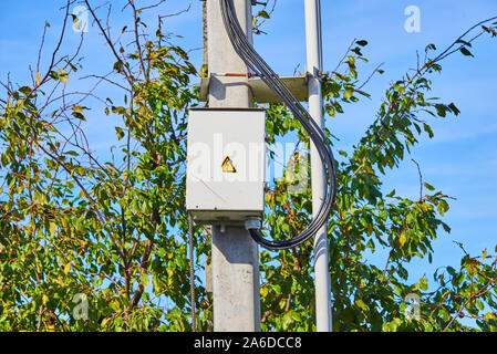 Locked rusty grey electric panel with hazard sign on the grey concrete pillar near trees and grey metal pipes Stock Photo