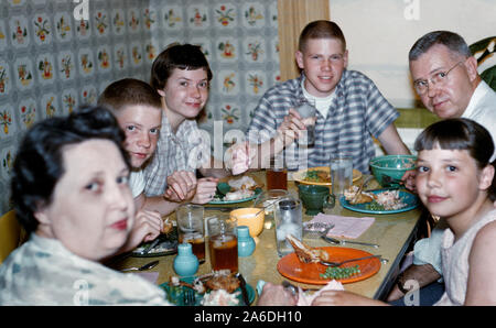 A 1950s Midwestern American family of six gather closely together for dinner around a small Formica-topped table to eat fried chicken, mashed potatoes and green peas served on multicolored Fiesta dinnerware with glasses of iced tea and water. Also note cigarette ashtray on the table, short hair styles, and patterned wallpaper typically in fashion during that mid-20th Century era. Historical photo. Stock Photo