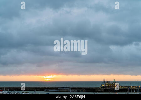 Sunrise over the English Channel, sea, and Ramsgate harbour in Kent, England. Grey storm clouds overhead with a thin band of orange sky on the horizon as the sun appears from behind clouds. Stock Photo