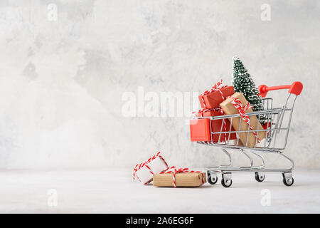 Shopping cart full of various gift boxes and a Christmas tree on light gray background. Christmas sale concept.