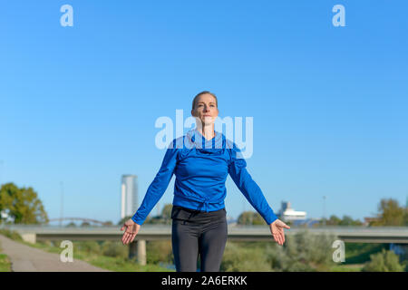 Sporty woman warming up with stretching exercises extending her arms before going jogging alongside an urban river on a cold sunny winter day in a low Stock Photo