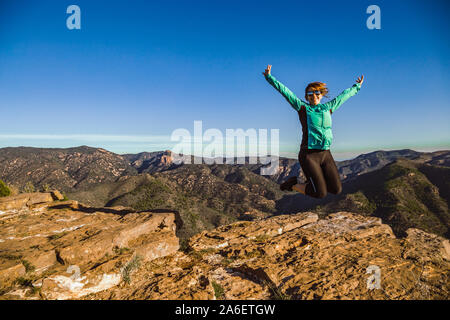 A young sporty woman jumping in the air on the rocky ground of a mountain with her arms raised to the blue sky - Hiker girl wearing sunglasses and leg Stock Photo