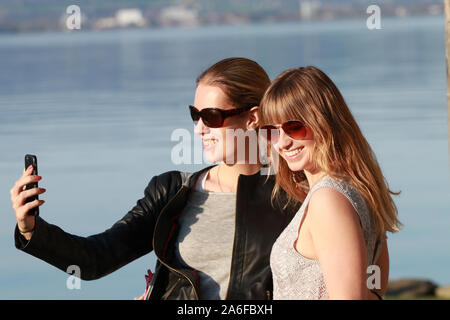 two girls with sunglasses by the lake taking selfies and having fun Stock Photo