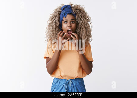 Shocked stupified african-american blond curly woman in stylish outfit, headband, gasping alarmed, drop jaw from surprised and fear, hold hands near Stock Photo