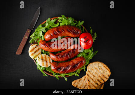 Grilled pork sausages with arugula and vegetables on black background. Close up, overhaed shot. Stock Photo