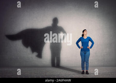 Brave woman keeps arms on hips, smiling confident, casting a superhero with cape shadow on the wall. Ambition and business success concept. Leadership Stock Photo