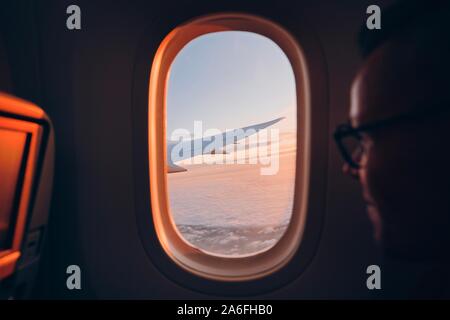 Man looking throught vindow of airplane during flight above clouds at sunset. Selective focus on wing. Stock Photo