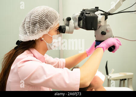 Young woman dentist doctor looks through a professional microscope in a dental clinic. A doctor in a disposable medical mask and cap. Advanced equipme Stock Photo