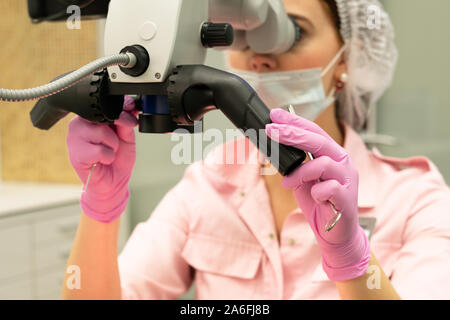 Young woman dentist doctor looks through a professional microscope in a dental clinic. A doctor in a disposable medical mask and cap. Advanced equipme Stock Photo