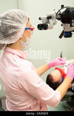Young female dentist looks through a professional microscope in a dental clinic. Doctor uses disposable gloves, a mask and a hat. Dentist works in the Stock Photo
