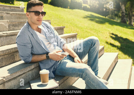 Outdoors Leisure. Stylish guy student in sunglasses wearing watch sitting on stairs in park with cup of coffee and notebook holding smartphone looking Stock Photo