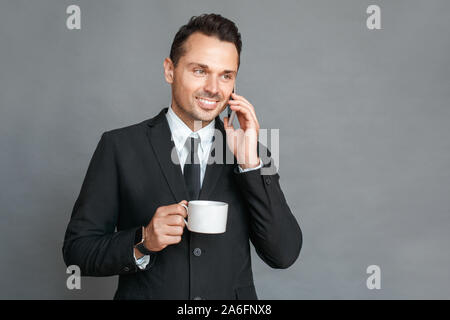 Businessman studio standing isolated on grey drinking coffee talking on smartphone smiling happy Stock Photo