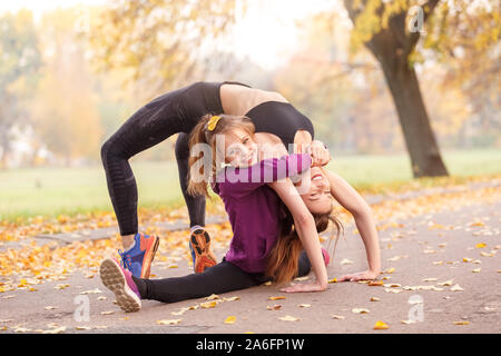 Outdoors leisure. Little girl doing splits while older doing arch exercise hugging in the autumn park smiling happy Stock Photo