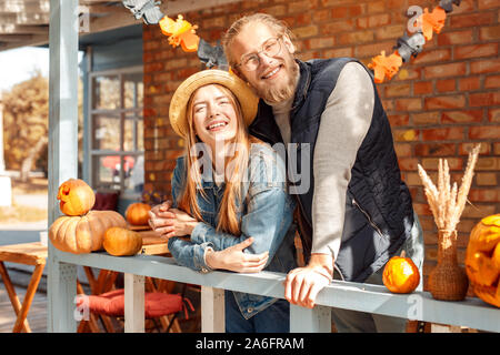 Halloween Preparaton Concept. Young couple leaning on fence at decorated home with jack-o'-lantern looking aside dreamful Stock Photo