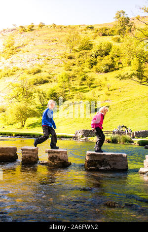 A little boy and girl hoping, jumping over the Dovedale stepping stones across a river, stream in the Peak District National Park, Derbyshire, Autism Stock Photo