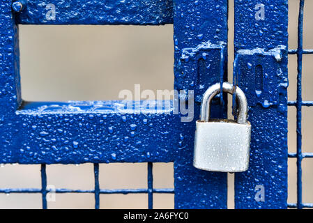 A blue metallic fence with a silver stainless steel padlock, security gate soaked with rain Stock Photo