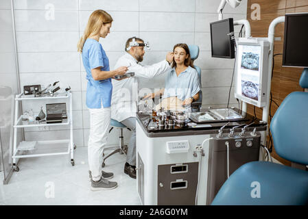 Senior otolaryngologist examining ear of a young patient with female assistant in ENT office, nurse giving medical tools Stock Photo