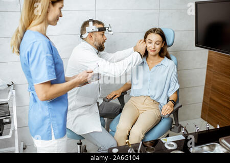Senior otolaryngologist examining ear of a young patient with female assistant in ENT office, nurse giving medical tools Stock Photo