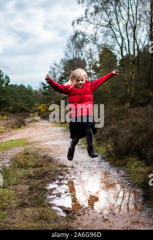 A little girl walks, runs and jumps through and into very muddy puddles in the countryside on a family walk wearing a warm red body warmer Stock Photo