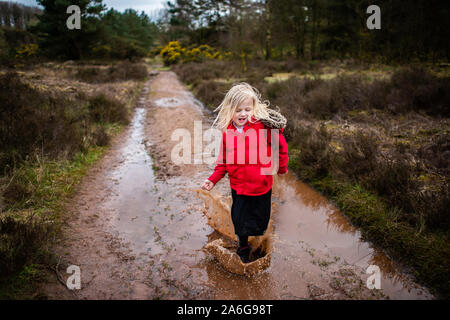 A little girl walks, runs and jumps through and into very muddy puddles in the countryside on a family walk wearing a warm red body warmer Stock Photo