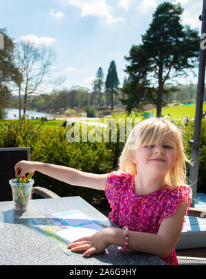 A cute little girl wearing a pretty dress enjoys doing some colouring while waiting for her dinner to arrive, dining outdoors in the sunshine Stock Photo