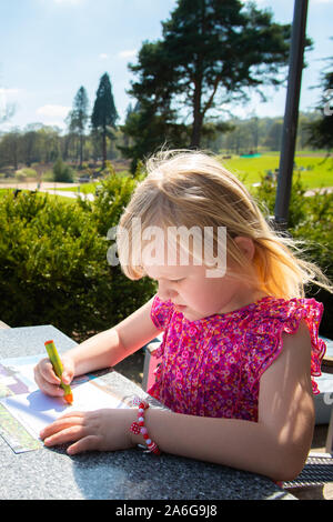 A cute little girl wearing a pretty dress enjoys doing some colouring while waiting for her dinner to arrive, dining outdoors in the sunshine Stock Photo