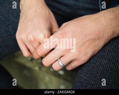Young male 20-25 years isolated hands with ring on left ring finger, sitting with hands between legs. No face visible. Stock Photo
