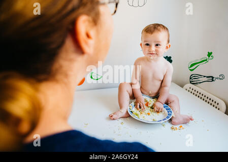 Baby smiling happily at his mother looking at him while eating with his hands in the kitchen. Stock Photo