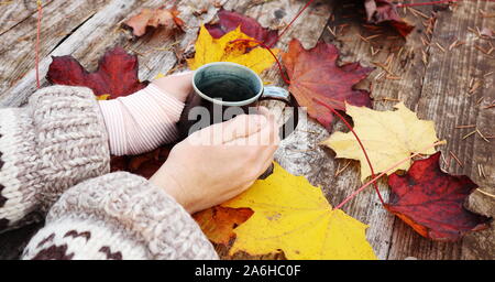 two hands holding a mug on a table scattered with colourful autumn maple leaves, left wrist wrapped in a compression bandage.  oversize wool sweater Stock Photo