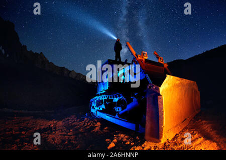 Man on bulldozer with head lamp under Milky Way view at night starry sky in the mountains Stock Photo