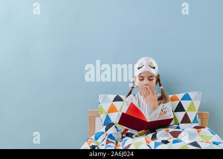 Yawning little girl is reading in bed in pajamas before going to sleep over blue