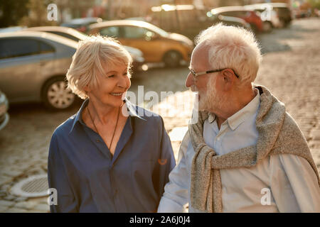 Portrait of happy elderly couple holding hands and looking to each other with smile while walking together through the street Stock Photo