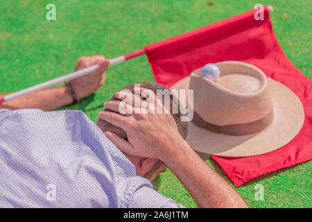 A man lying on a green , with his hand on his head, next to a red flag, a hat and a golf ball on top. Concept of a day of relaxation and leisure. Colo Stock Photo