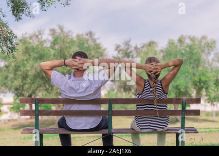 A woman with blond hair relaxed next to her boyfriend, stretching her hands over her head, sitting on a wooden stool in the park. Concept of a day of Stock Photo