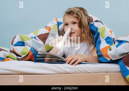 Adorable little girl reading in bed with flashlight under blanket Stock Photo